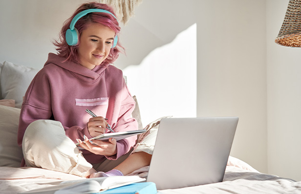 young woman wearing headphones studying at home with laptop and notebook in cozy bedroom learning environment focused on tasks related to four essential skills