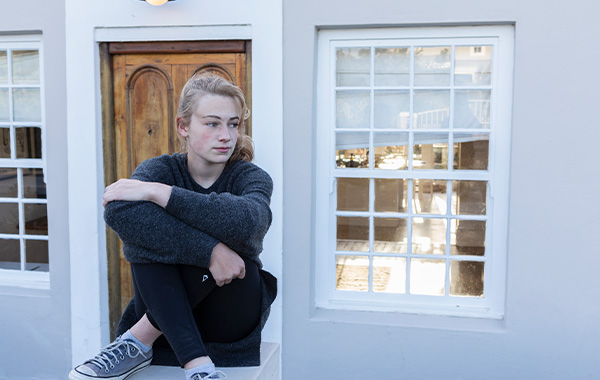 person sitting by window wearing gray sweater and black leggings with sneakers looking contemplative in a two tone setting